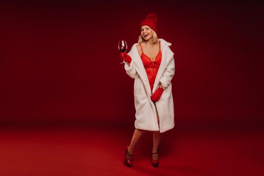 portrait of a smiling girl in a white fur coat and underwear holding a glass of champagne on a red background.