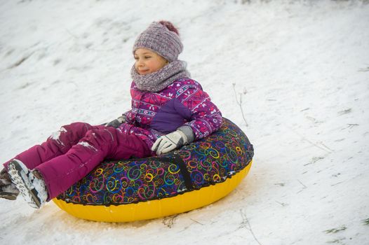 a little girl in winter in purple clothes and an inflatable circle rides down the hill on the street.
