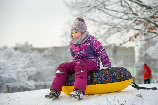 a little girl in winter in purple clothes and an inflatable circle rides down the hill on the street.