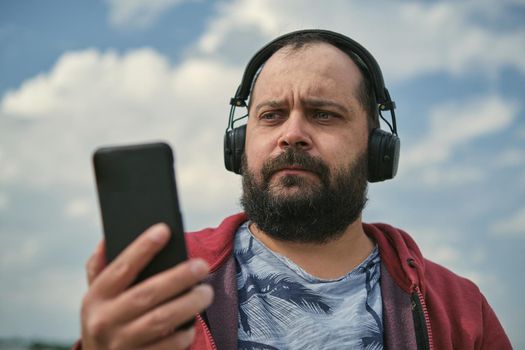 Middle-aged European man in headphones outdoors listening to music against the background of the sky, mobile phone in his hand