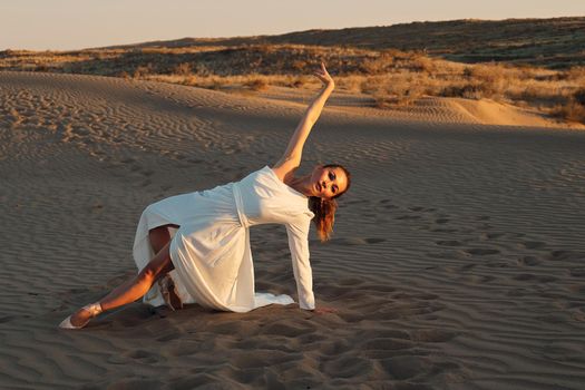 A girl in a fly white dress dances and poses in the sand desert at sunset.