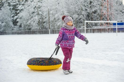 a little girl in winter in purple clothes and an inflatable circle walks on the street in a snow-covered forest.