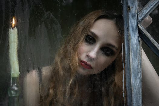 Beautiful sad young girl looking out the window with a candle, good for book cover