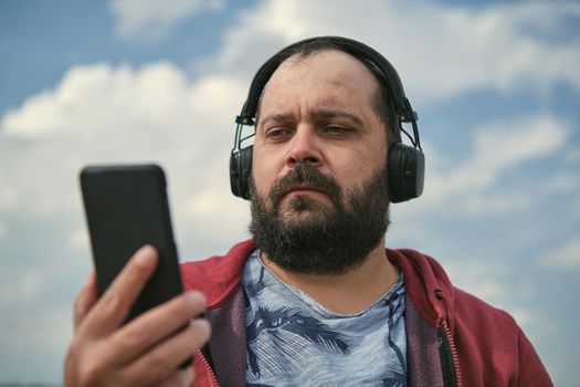 Middle-aged European man in headphones outdoors listening to music against the background of the sky, mobile phone in his hand