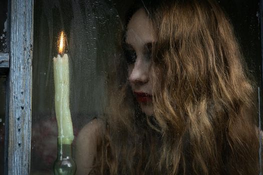 Beautiful sad young girl looking out the window with a candle, good for book cover