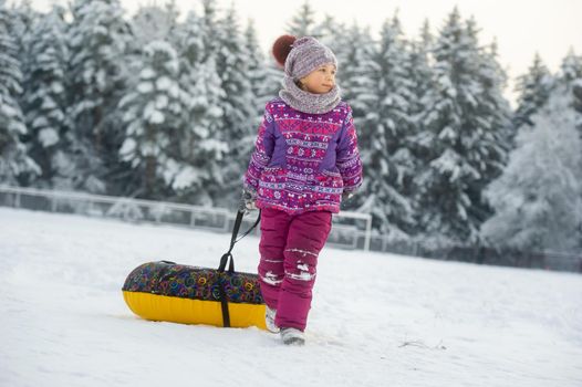 a little girl in winter in purple clothes and an inflatable circle walks on the street in a snow-covered forest.