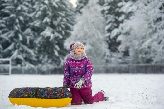 a little girl in winter in purple clothes and an inflatable circle walks on the street in a snow-covered forest.