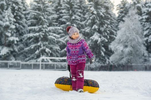 a little girl in winter in purple clothes and an inflatable circle walks on the street in a snow-covered forest.