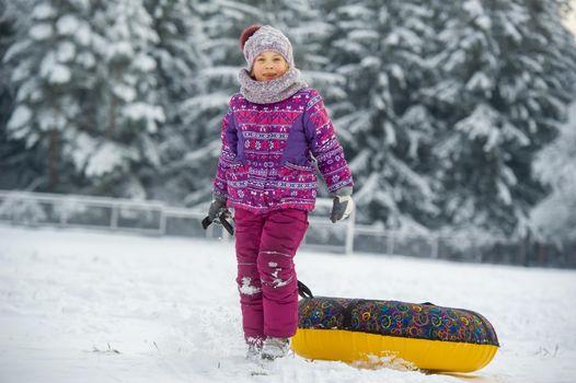 a little girl in winter in purple clothes and an inflatable circle walks on the street in a snow-covered forest.