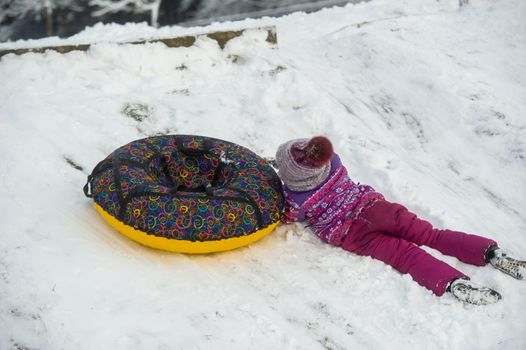 a little girl in winter in purple clothes and an inflatable circle rides down the hill on the street.