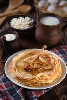 Homemade thin pancakes with honey stacked in a stack, on a wooden table with a mug of milk, a pot of sour cream and eggs in a basket. Traditional Slavonian, pagan holiday (Maslinitsa). Country style food.