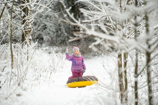 a little girl in winter in purple clothes and an inflatable circle walks on the street in a snow-covered forest.