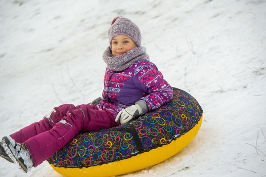 a little girl in winter in purple clothes and an inflatable circle rides down the hill on the street.