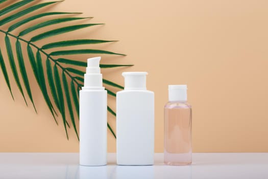 Set of white unbranded cosmetic bottles for skin care in a row on white table against bright beige background with palm leaf. Concept of organic, natural, eco friendly cosmetics and beauty products