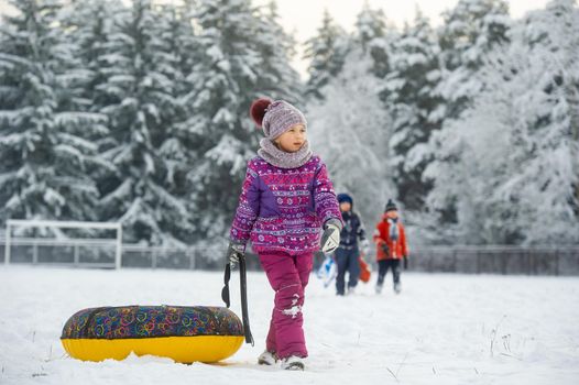 a little girl in winter in purple clothes and an inflatable circle walks on the street in a snow-covered forest.