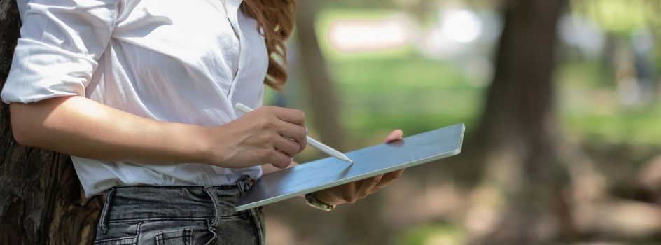 Close up hand of woman with stylus pen writing on digital notepad, touching on digital tablet screen working in park.