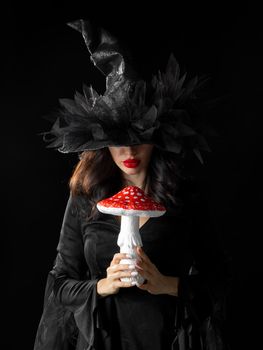 Portrait of sorceress witch woman in halloween costume holding poisonous mushroom for potion isolated on black background
