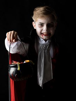 Boy with cauldron basket and worm sweets dressed as vampire for Halloween party going trick or treating. Studio portrait isolated over black background