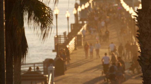 People walking, wooden pier in California USA. Oceanside waterfront vacations tourist resort. Ocean beach summertime sunset atmosphere. Blurred crowd strolling seaside boardwalk. Defocused palm trees.