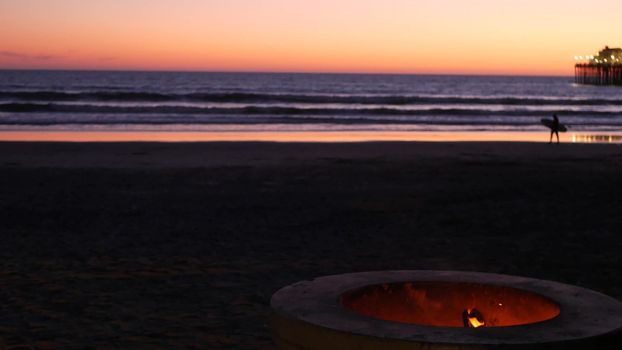 Campfire pit by Oceanside pier, California USA. Camp fire burning on ocean beach, bonfire flame in cement ring place for bbq, sea water waves. Romantic evening twilight dusk sky. Surfer with surfboard