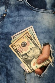 Hand holds several different value US dollar paper currency banknotes in jeans rip hole, low angle view