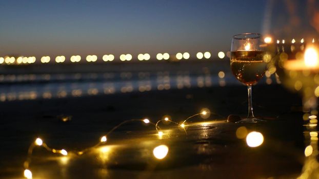 Candle flame lights in glass, romantic beach date, California ocean waves, sea water. Candlelight seamless looped cinemagraph. Wineglass on sand, garland in twilight dusk. Illuminated pier reflection.