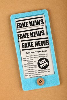 Close up felt craft blue smartphone with FAKE NEWS newspapers feed from screen over brown paper background, elevated top view, directly above