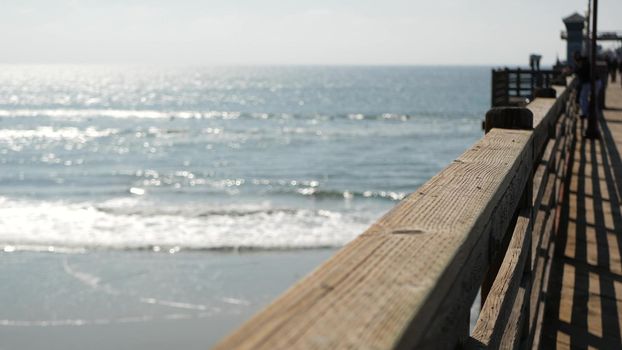 Railings of old wooden pier, people walking on waterfront boardwalk, Oceanside beach atmosphere, California coast USA. Defocused seascape, pacificocean water waves. Los Angeles summertime vacations.