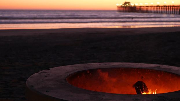 Campfire pit by Oceanside pier, California USA. Camp fire burning on ocean beach, bonfire flame in cement ring place for bbq, sea water waves. Romantic evening twilight sky, dusk after summer sunset.