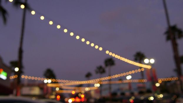 Defocused illuminated electric garland, palm trees silhouettes, Oceanside California USA. Ocean beach tropical pink sunset, pacific coast purple twilight sky. Los Angeles vibes. Bulb lights glowing.