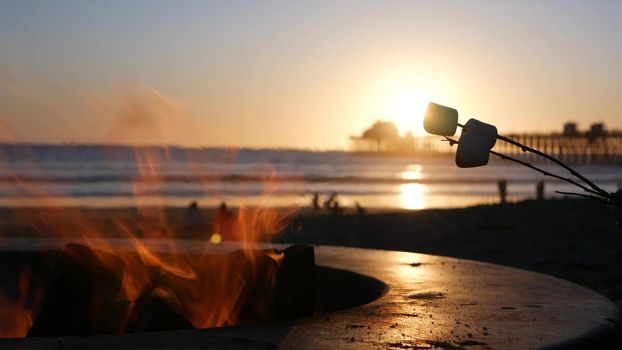 Campfire pit by Oceanside pier, California USA. Camp fire on ocean beach, bonfire flame in cement ring place for bbq, sea water waves. Heating, roast or toast marshmallow on stick. Romantic sunset sky