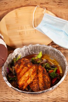 Grilled chicken in a takeout container with grilled broccoli and onions with a football, a wooden football, and a paper mask in background