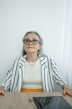 Senior grey haired businesswoman in striped jacket with eyeglasses is working in her office sitting at the desk. Happy retirement, employment and labor.