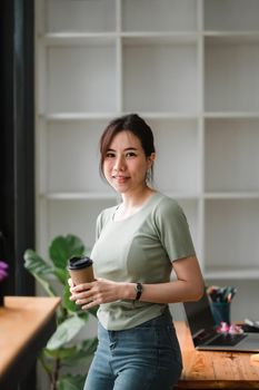 Asian woman standing on front of office workplace while hand holding a cup of coffee and looking outside.