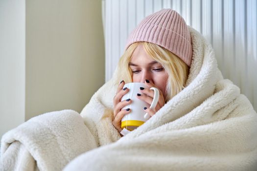 Heating during the cold winter autumn season. Young female teenager woman warming herself with a blanket hat, a central heating radiator, hot tea