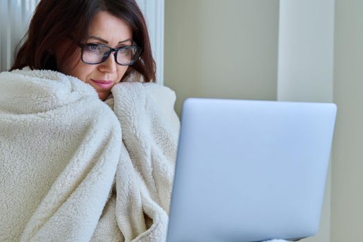 A frozen mature woman working at home with a laptop, warming herself with a blanket and heating radiator. Autumn winter cold season