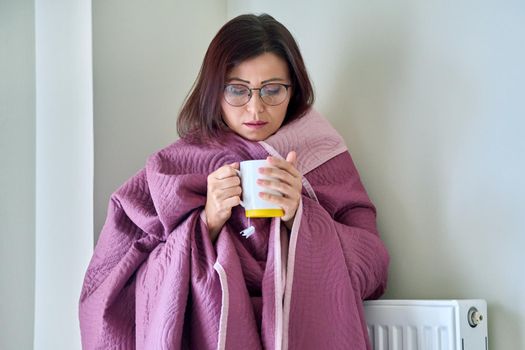 Heating during the cold winter autumn season. An adult woman warming herself with a blanket, a central heating radiator, hot tea.