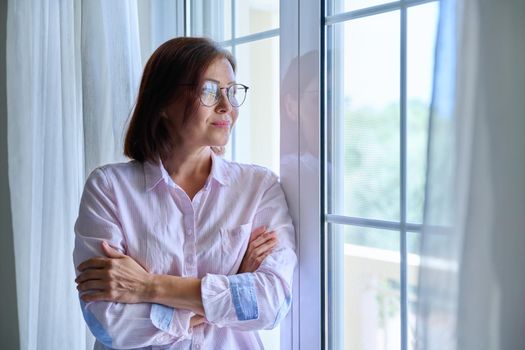 Portrait of a mature confident woman looking out the window. Middle-aged female wearing glasses with crossed arms, calm, smiling, resting