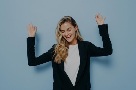 Portrait of carefree happy woman with blonde dyed hair raising her arms and dancing with closed eyes and smile on face while standing isolated against blue background. Happiness concept