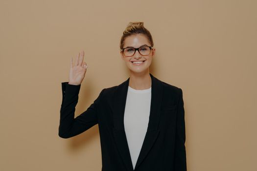 Positive young caucasian woman in classic wear isolated on beige background showing ok sign or okay gesure with fingers and smiling at camera, female wearing eyeglasses saying that everything fine