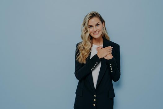 Young lovely woman with blond hair in black blazer expressing love and admiration while holding both hands on chest and smiling happily at camera, standing isolated over blue wall with copy space