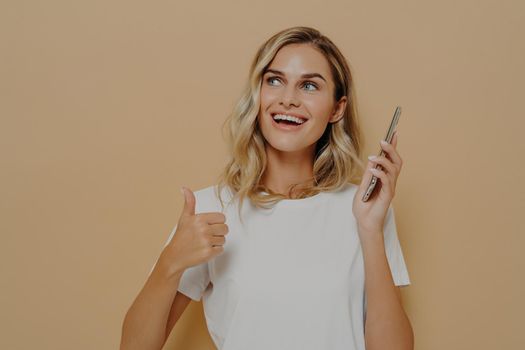 Young satisfied girl in casual clothes holding mobile phone and showing thumb up gesture, being excited to hear positive news while standing isolated over brown background, looking upside with smile