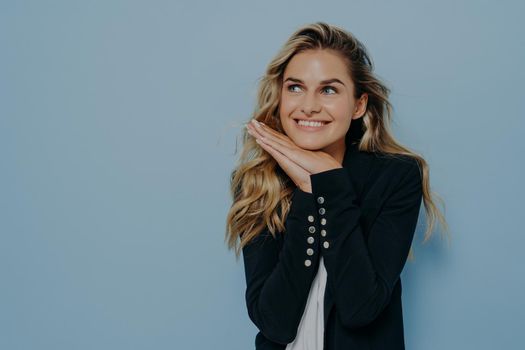 Delightful young european woman with blonde wavy hair wearing black blazer over white tshirt feeling happy of hearing compliment from friends, smiling and looking upside. Positive human emotions