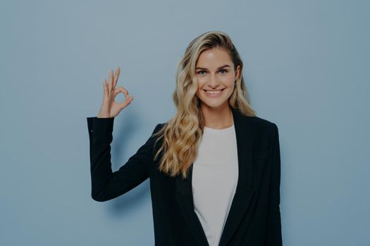 Cheerful blonde female student in casual clothes demonstrating agreement while showing okay gesture and looking at camera with positive face expression. Body language concept