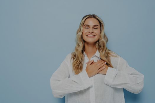 Pleased happy thankful woman with blond wavy hair in white oversize shirt gesturing appreciation and love and gratitude by holding hands together on chest, eyes closed, isolated over blue wall