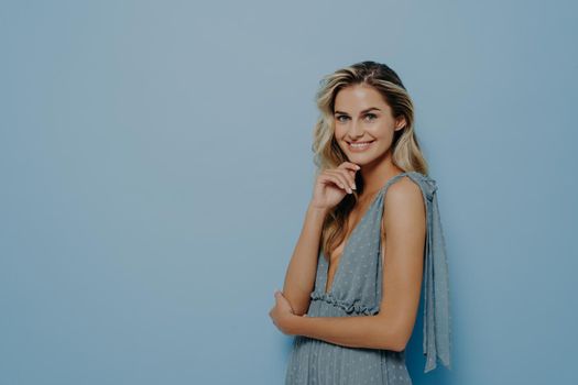 Fashion studio photo of beautiful woman with blond wavy hair and natural makeup, wears delicate party dress, posing sideways with folded hands and gently touching her face with fingers, softly smiling
