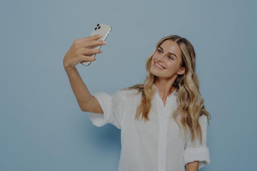 Beautiful caucasian young female with long blonde dyed hair holding mobile phone and posing for selfie, looking at screen with flirty smile, attractive woman making photo for social networks online