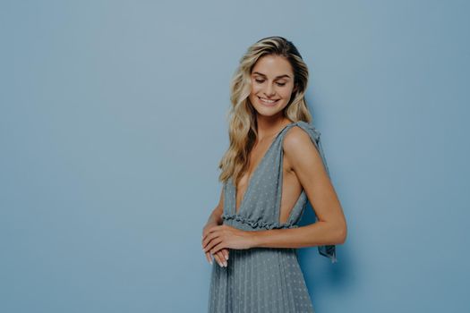 Studio portrait of young smiling beautiful female model with long blonde wavy hair in soft blue evening dress with deep neckline posing sideways on pastel blue background with copy space
