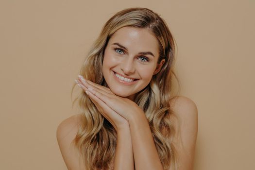 Smiling merry blonde young woman with long wavy hair feeling happy and showing it with gesture, resting head on hands together under chin, isolated in front of orange background