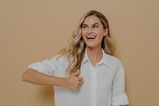 Happy blonde girl in white shirt showing thumbs up gesture to someone while looking sideways, enjoying moment and smiling with wide toothy smile while standing isolated next to orange wall
